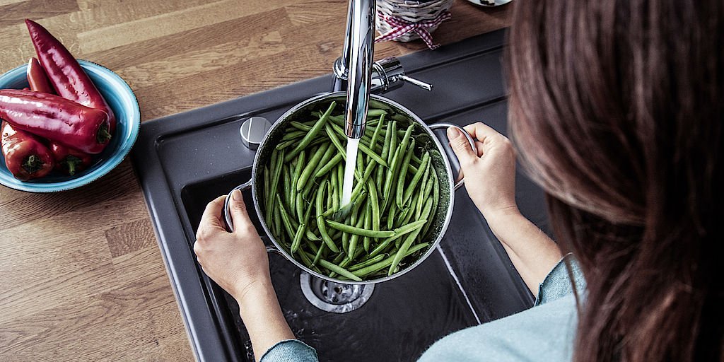 Eine Frau haelt ein Topf mit Bohnen unter der laufenden Kuechenspuehle