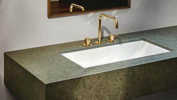 Ein elegantes Waschbecken aus weißem Keramik, umgeben von einer grünen Steinplatte und goldenen Wasserhähnen.
